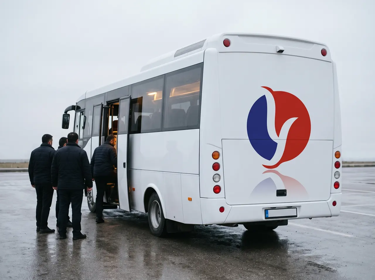 Yugam Grup logolu beyaz servis otobüsü, yağmur sonrası ıslak otoparkta; personel açık kapıdan araca biniyor